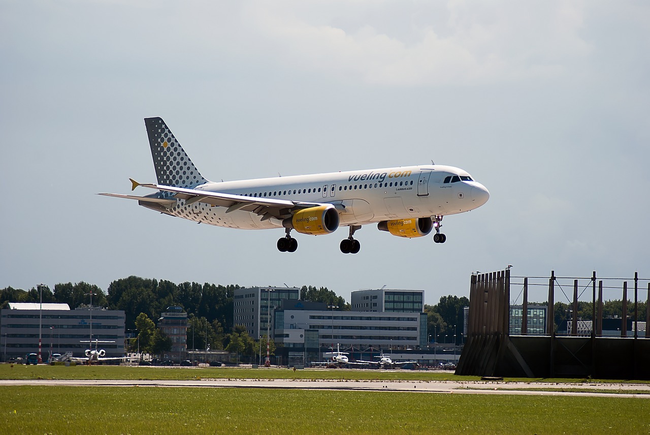 A Vueling aircraft mid-flight, descending onto the runway. Buildings can be seen in the background.