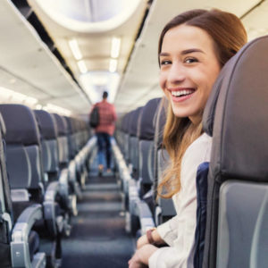 Young woman smiles while waiting to exit a commercial flight. A male passenger is exiting in the background.