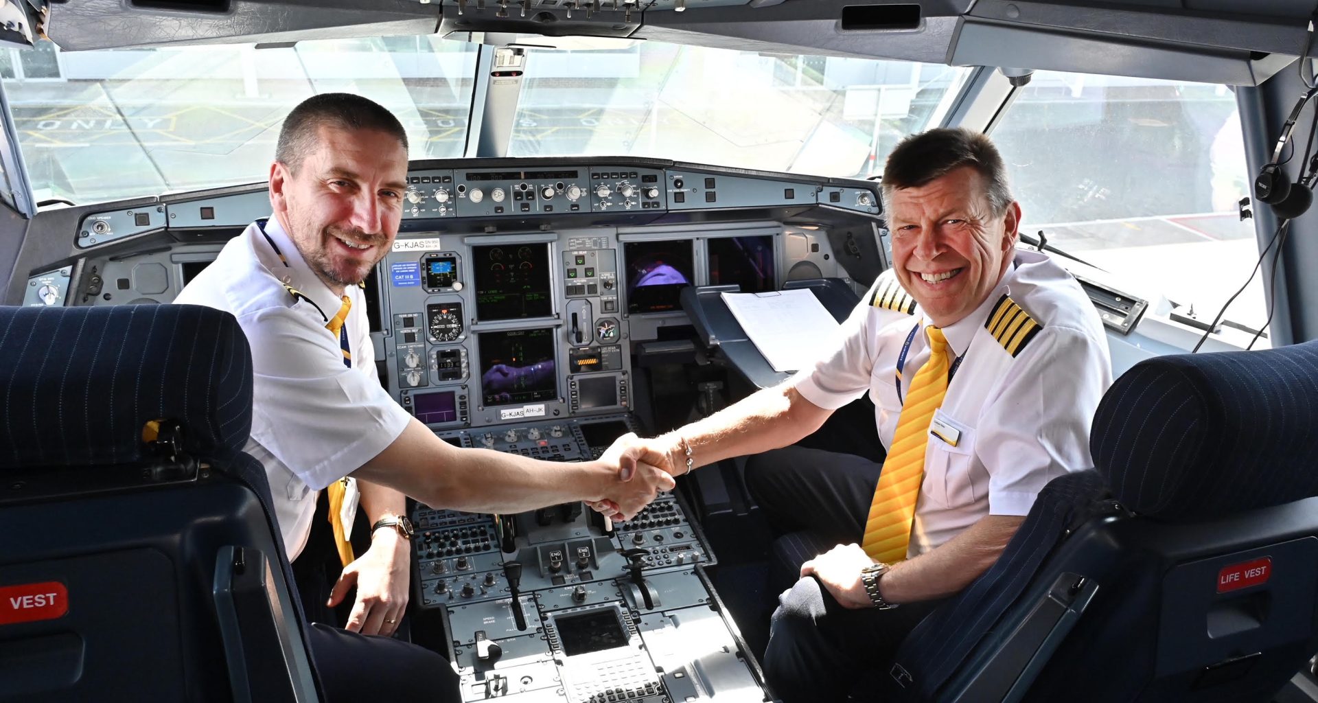 Two male pilots in cockpit look into camera, shaking hands.