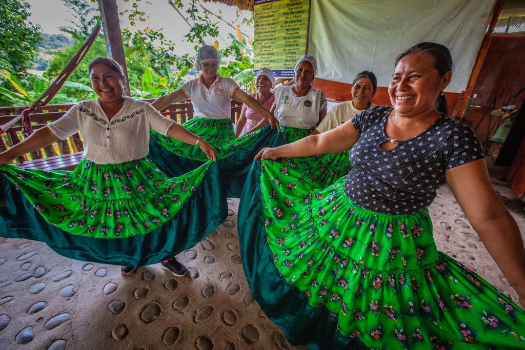 48138690752 affa3d8557 k e1666788389695 Las mujeres indígenas se paran con faldas anchas tiradas hacia los lados dentro de una habitación de madera.