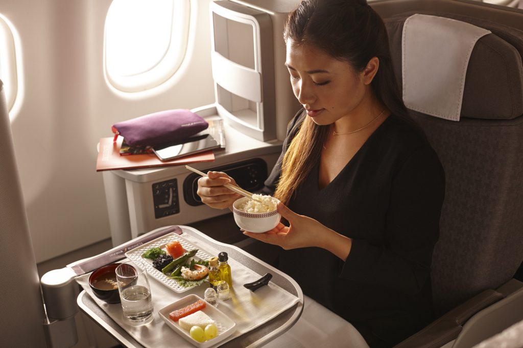 iberia 4751091420053885 e1666776446248 Mujer sentada en clase ejecutiva en un vuelo. Una bandeja de comida está al frente y ella sostiene un tazón pequeño del que está comiendo.