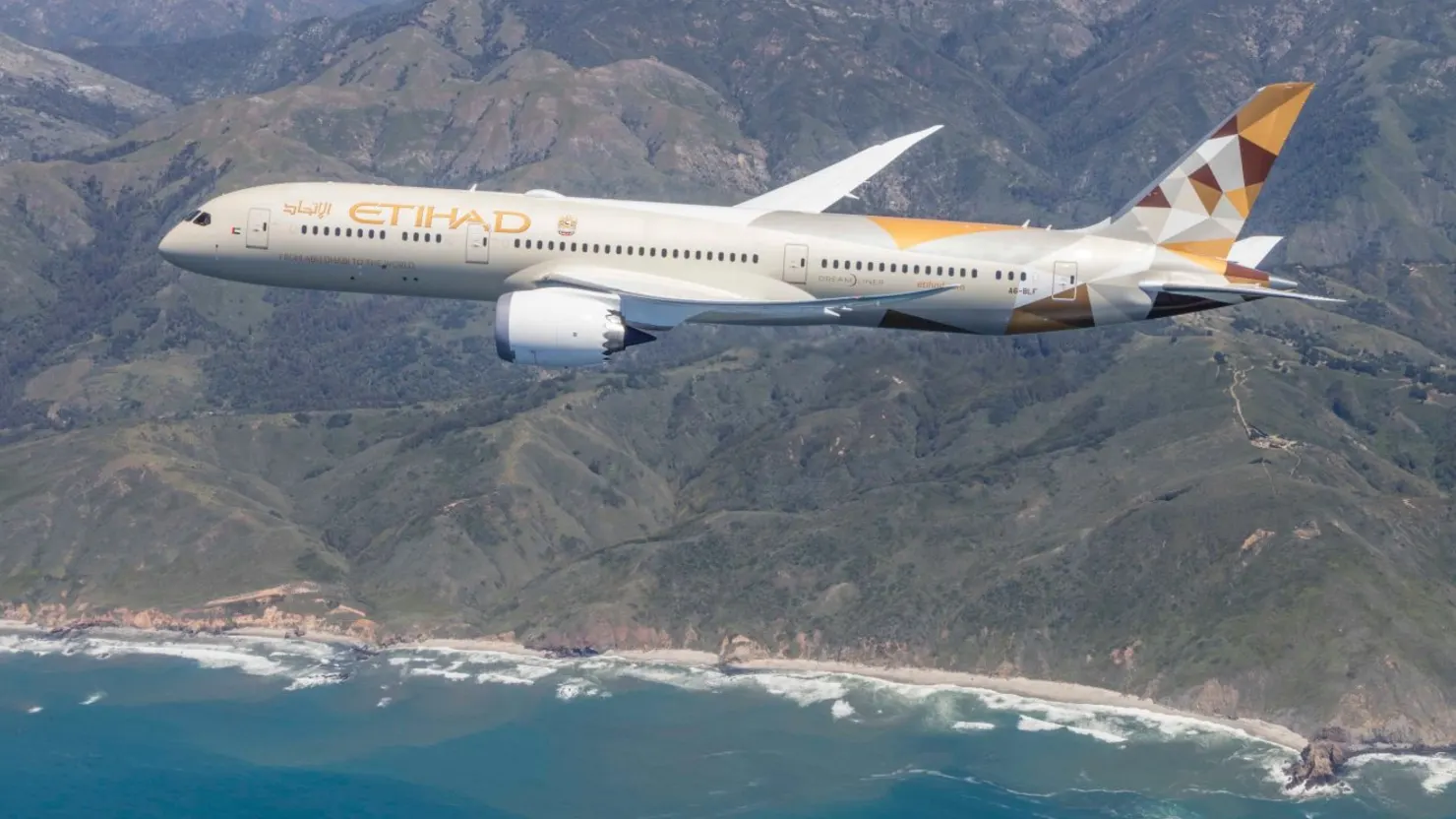 An Etihad Airways Boeing 787-9 Dreamliner flys above a scene in view of mountains and an ocean