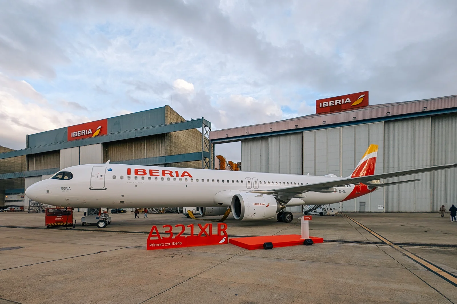 Image shows a white and red and orange Iberia Airbus A321XLR parked in front of an Iberia branded hangar on a cloudy day 