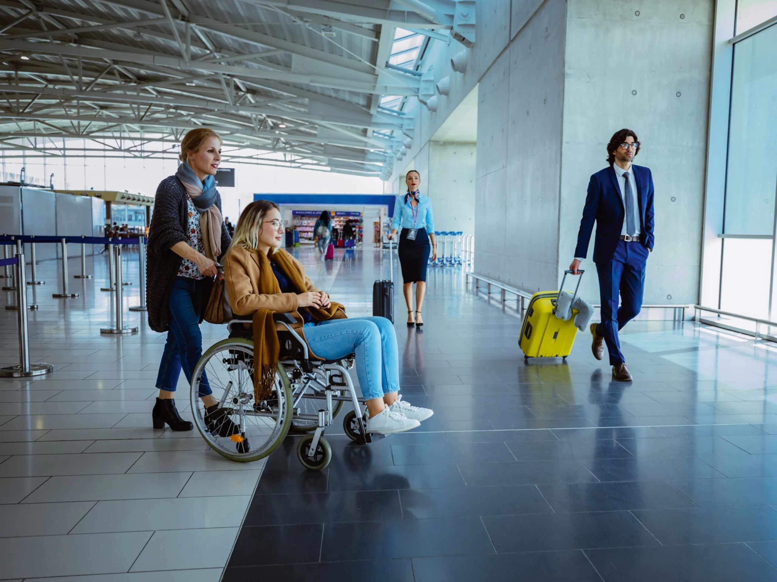 Image shows a woman pushing another woman in a wheelchair at an airport during the daytime. A passenger and aircraft crew member are also visible pulling their suitcases