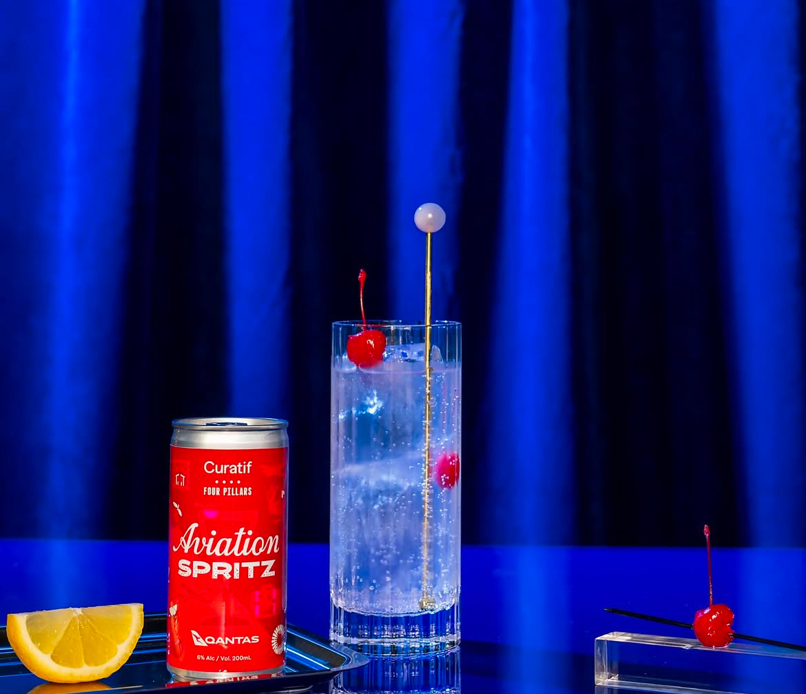 The Aviation Spritz can in front of a blue background with the drink in a glass beside it. As well as decorative cherries and lemons scattered across