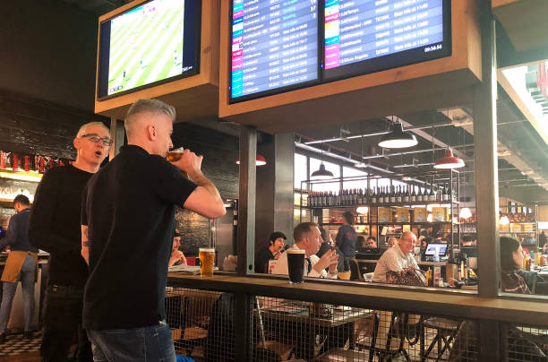 Two men drink at an airport bar in front of two boarding screens.