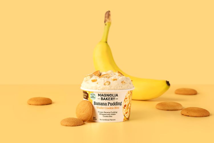 A promotional shot of Magnolia Bakery’s frozen banana pudding. The pudding container has no lid and reveals the pudding, there are cookies surrounding the container as well as a banana behind it. 