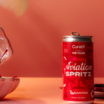 The Aviation Spritz can in front of a pastel pink background. A glass with the drink is beside the can with cherries in it, as well as a pink shell clam decoration with cherries inside it