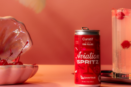 The Aviation Spritz can in front of a pastel pink background. A glass with the drink is beside the can with cherries in it, as well as a pink shell clam decoration with cherries inside it