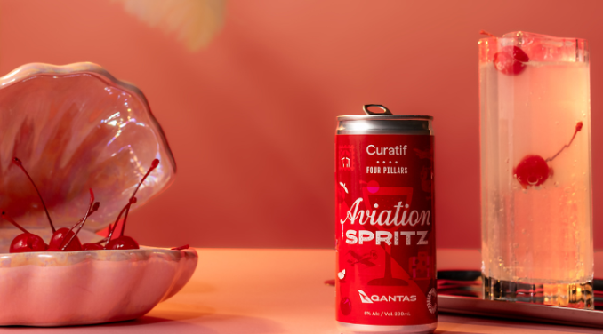 The Aviation Spritz can in front of a pastel pink background. A glass with the drink is beside the can with cherries in it, as well as a pink shell clam decoration with cherries inside it