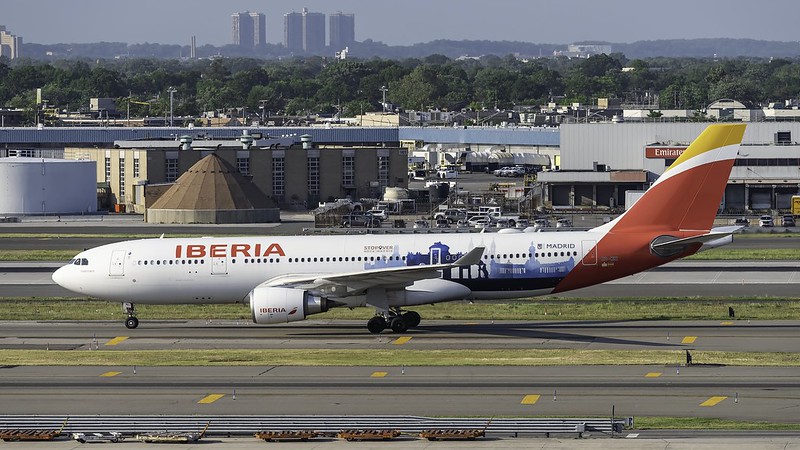 Image shows an Iberia Airlines aircraft on a runway during the day with airport buildings and vehicles visible in the background