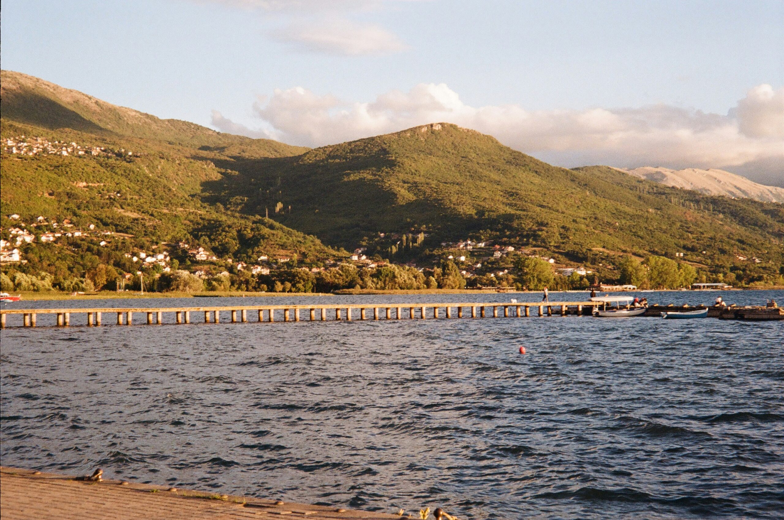 A wooden pier stretching into a lake with small boats docked nearby. The background features green mountains dotted with houses, and the scene is bathed in warm sunlight. The view of the Ohrid lake.