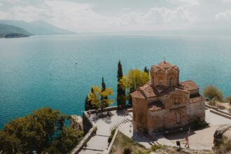 A historic stone church with a red-tiled roof sits on a cliff overlooking a vast, tranquil blue lake. The surrounding landscape includes lush green trees, a winding stone pathway, and small boats floating on the water. Mountains stretch across the horizon under a partly cloudy sky.