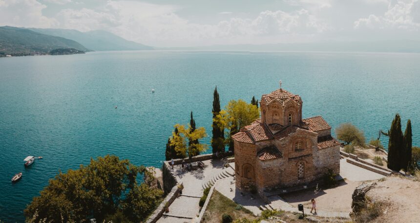 A historic stone church with a red-tiled roof sits on a cliff overlooking a vast, tranquil blue lake. The surrounding landscape includes lush green trees, a winding stone pathway, and small boats floating on the water. Mountains stretch across the horizon under a partly cloudy sky.