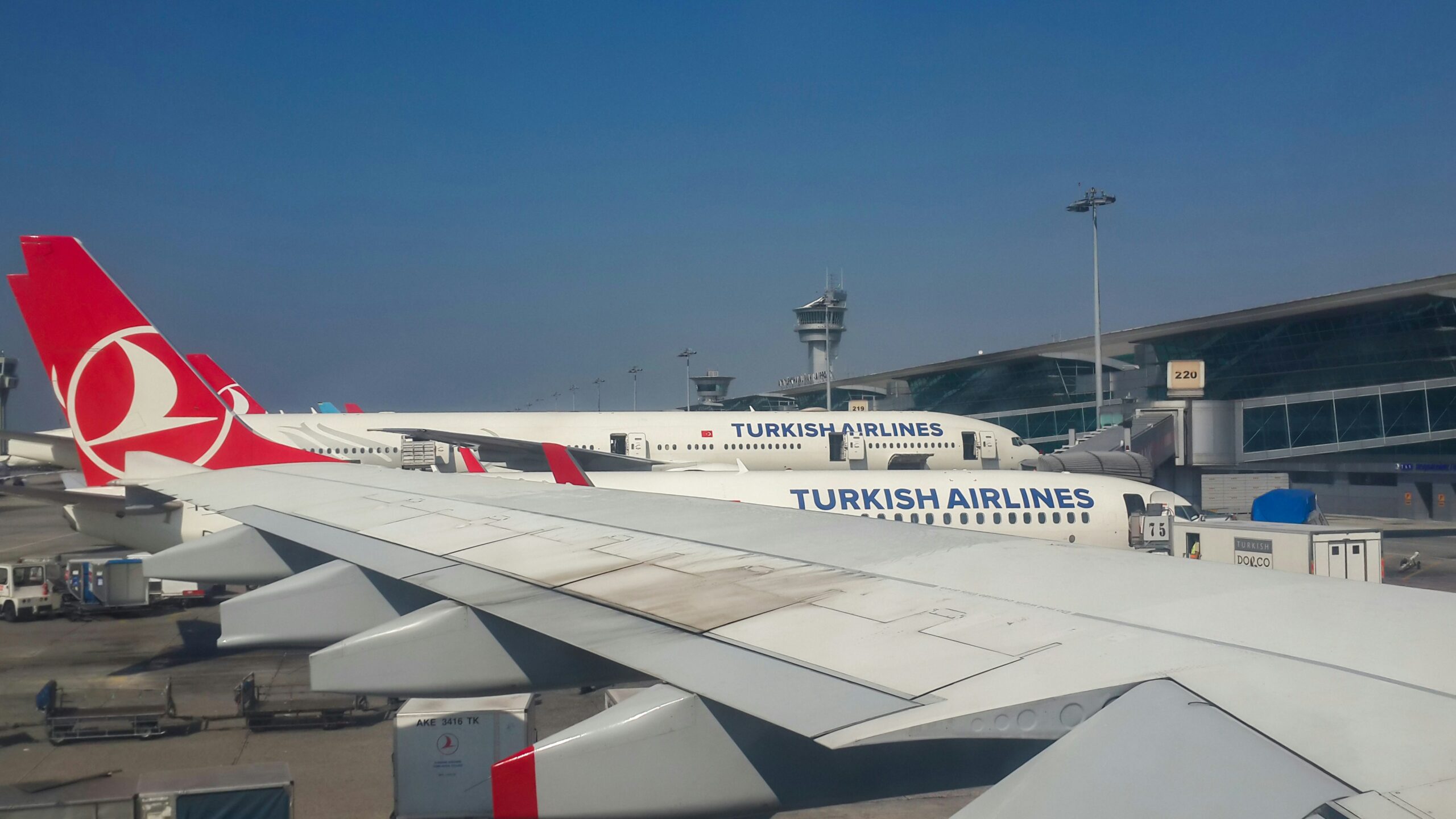 A view from an airplane window showing multiple Turkish Airlines aircraft parked at an airport terminal. The red tail fins with the airline's logo are visible, along with the terminal building, a control tower, and various ground support equipment. The wing of the plane from which the photo was taken is also prominently in the foreground.