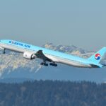 A Korean Air Boeing 777-300ER aircraft is captured mid-takeoff against a backdrop of snow-capped mountains and a clear blue sky. The airplane is painted in the airline’s signature light blue livery, featuring the "Korean Air" logo on the fuselage and the red-and-blue Taegeuk symbol on the tail. The landing gear is still in the process of retracting as the aircraft ascends. Below the plane, a dense forest and some industrial buildings with smoke stacks are visible.