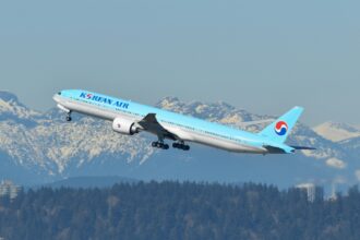 A Korean Air Boeing 777-300ER aircraft is captured mid-takeoff against a backdrop of snow-capped mountains and a clear blue sky. The airplane is painted in the airline’s signature light blue livery, featuring the "Korean Air" logo on the fuselage and the red-and-blue Taegeuk symbol on the tail. The landing gear is still in the process of retracting as the aircraft ascends. Below the plane, a dense forest and some industrial buildings with smoke stacks are visible.