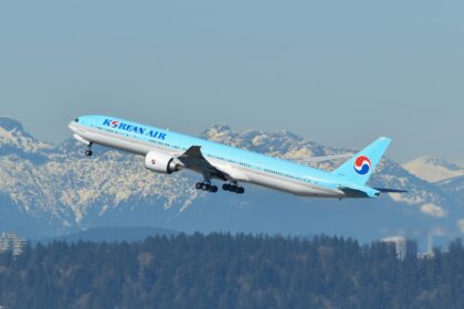 A Korean Air Boeing 777-300ER aircraft is captured mid-takeoff against a backdrop of snow-capped mountains and a clear blue sky. The airplane is painted in the airline’s signature light blue livery, featuring the "Korean Air" logo on the fuselage and the red-and-blue Taegeuk symbol on the tail. The landing gear is still in the process of retracting as the aircraft ascends. Below the plane, a dense forest and some industrial buildings with smoke stacks are visible.
