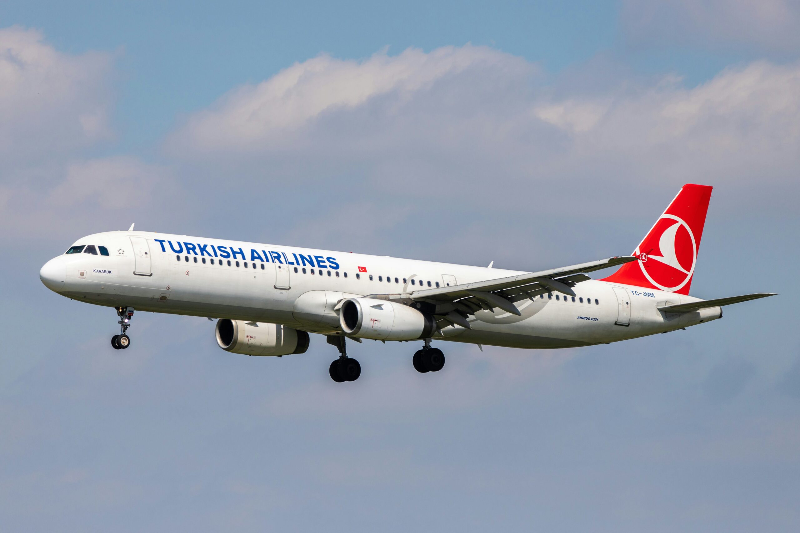 A Turkish Airlines airplane with a white fuselage and red tail fin, flying with landing gear extended. The airline’s name is visible on the side, along with the Turkish flag.