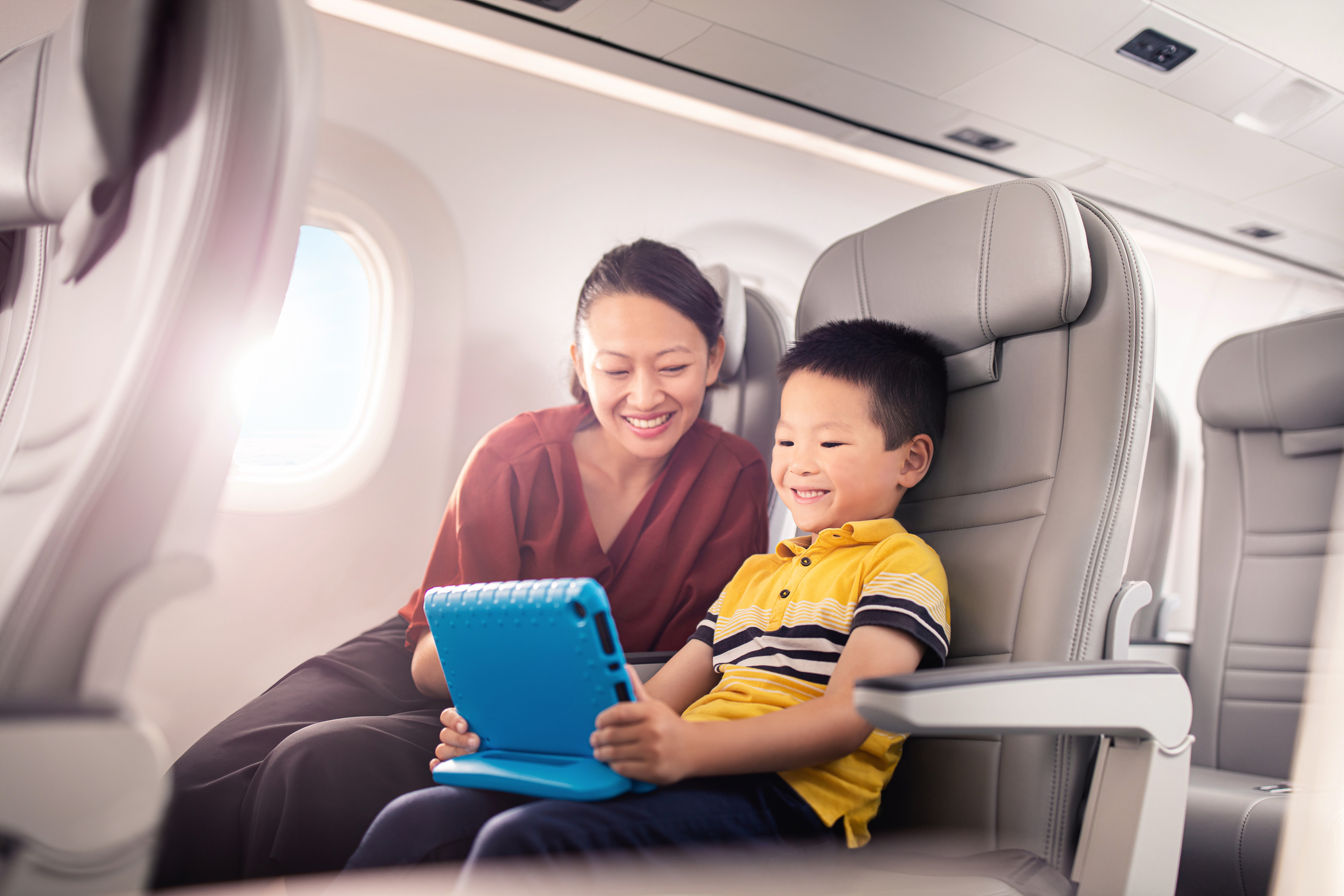 Mother and young boy sitting on an airplane, smiling while using a blue tablet, emphasizing in-flight entertainment and connectivity.