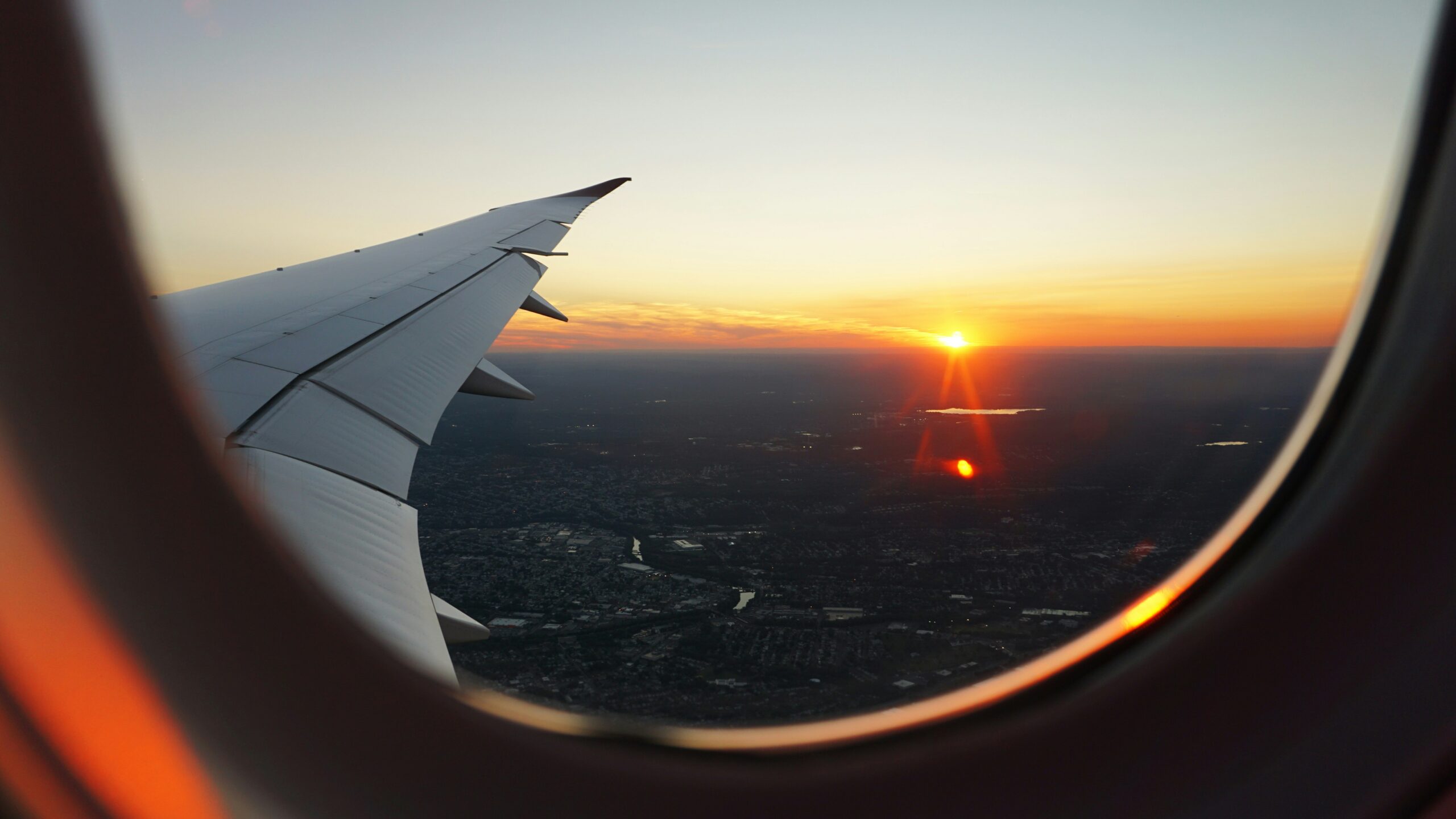 A view of a sunset from an airplane window, showing the aircraft wing and a city landscape far below.