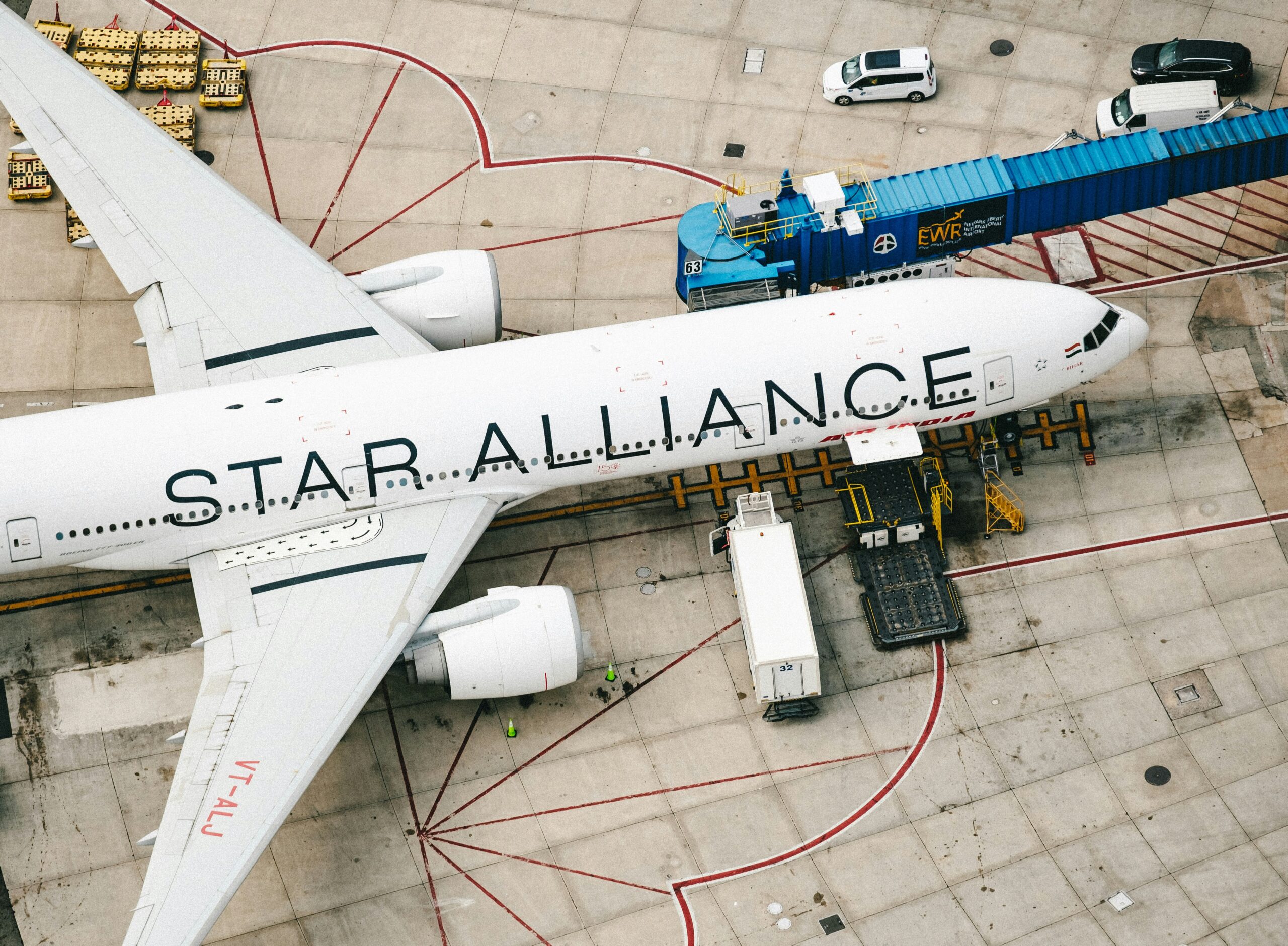 Aerial view of a Star Alliance airplane parked at an airport gate, with a passenger bridge, service vehicles, and ground crew visible.