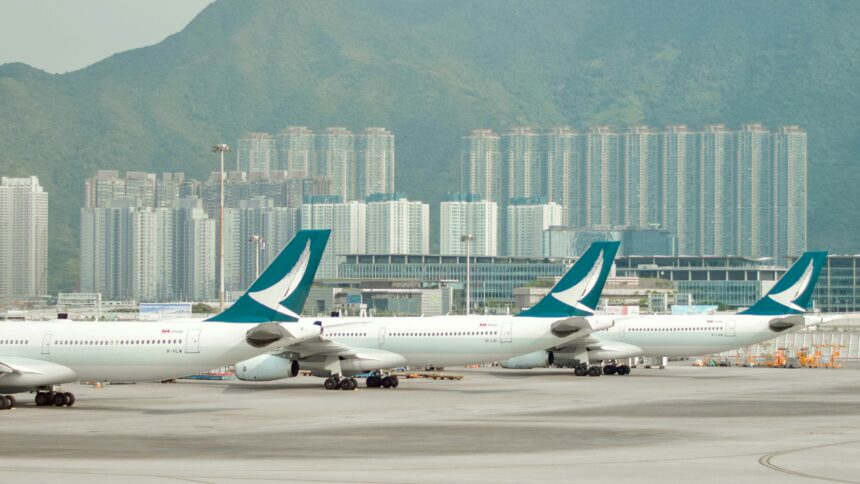 Multiple Cathay Pacific aircraft parked at Hong Kong International Airport, with city skyscrapers and green mountains in the background.