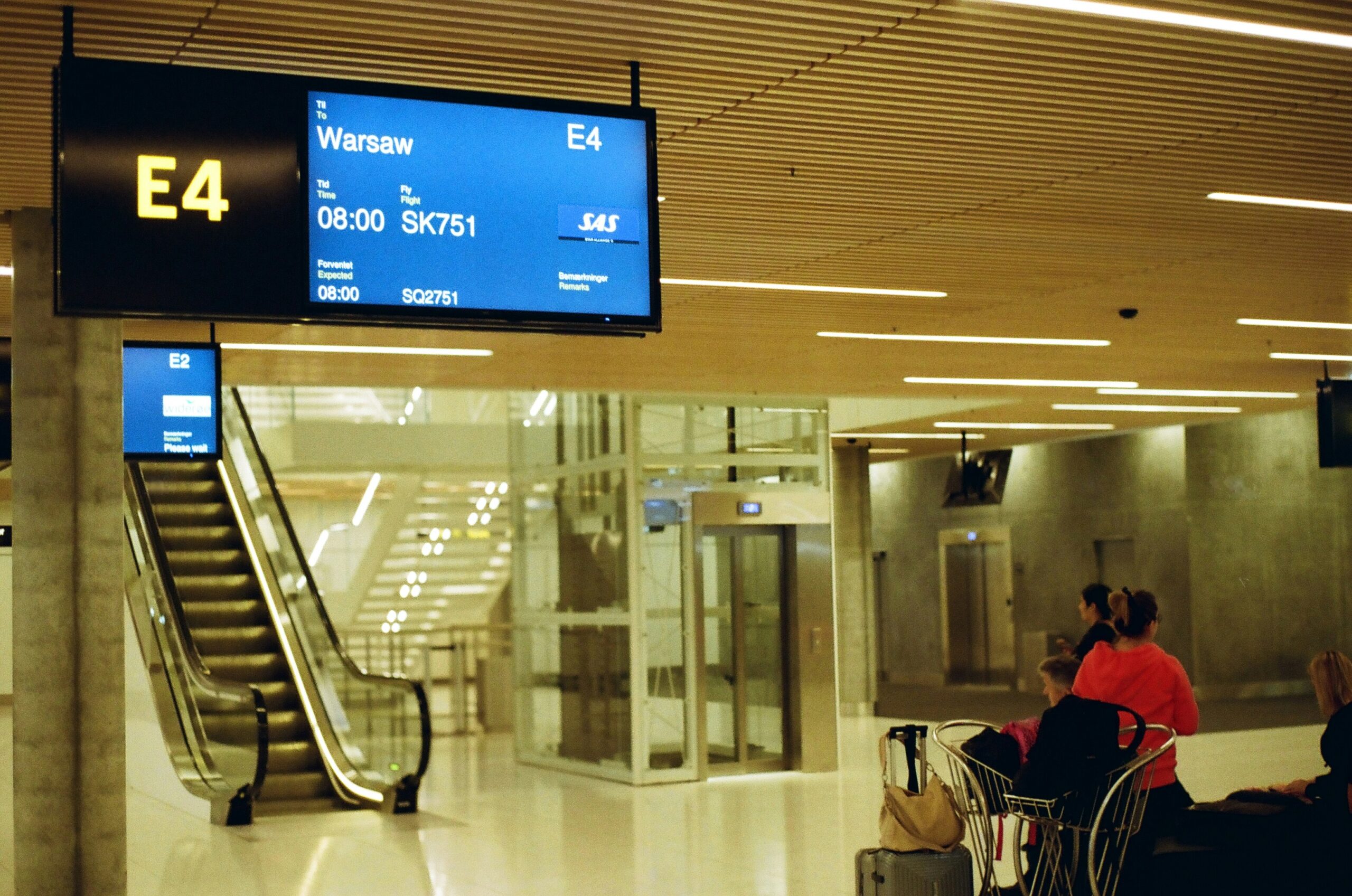 A modern airport terminal with a digital departure board displaying flight SK751 to Warsaw at gate E4. The terminal has an escalator, elevators, and passengers waiting with luggage.
