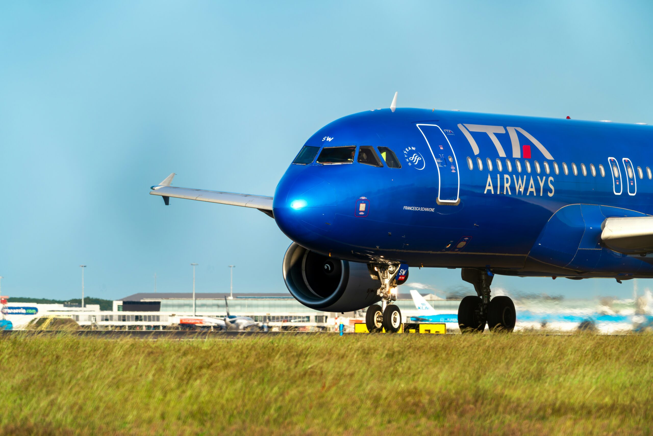 Blue ITA Airways plane taxiing on a runway, with airport buildings and other aircraft in the background under a clear sky.