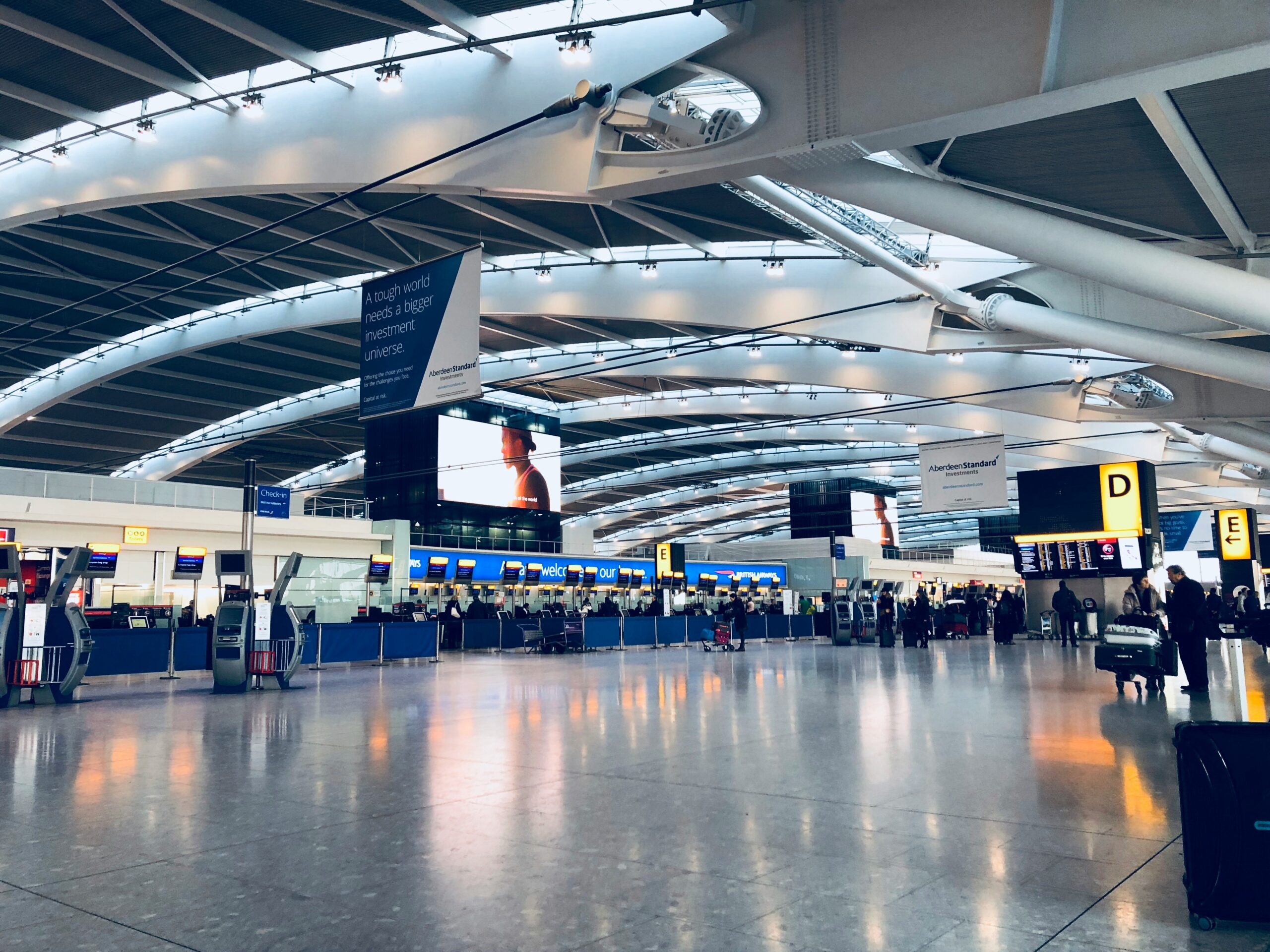 A spacious airport departure hall at London Heathrow Airport with check-in counters, passengers, and digital signboards under a modern curved ceiling.