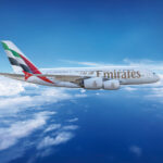 Emirates Airbus A380 flying in a clear blue sky above the clouds, featuring the airline's branding and UAE flag on the tail.