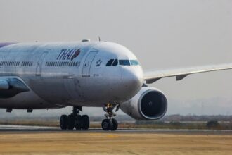 A Thai Airways airplane is taxiing on an airport runway, with its front landing gear and one engine visible.
