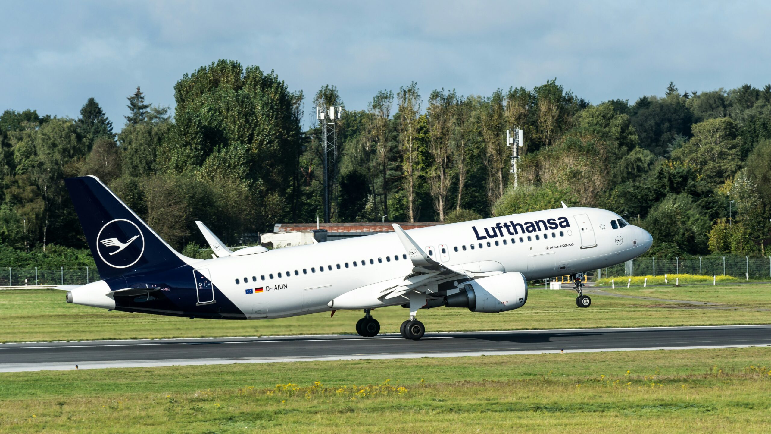 A Lufthansa aircraft in flight, departing from an airport runway, with a backdrop of clear skies and airport facilities.