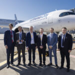 A group of airline and industry executives posing in front of a Lufthansa City Airlines Airbus A320neo on the airport tarmac during a delivery event.