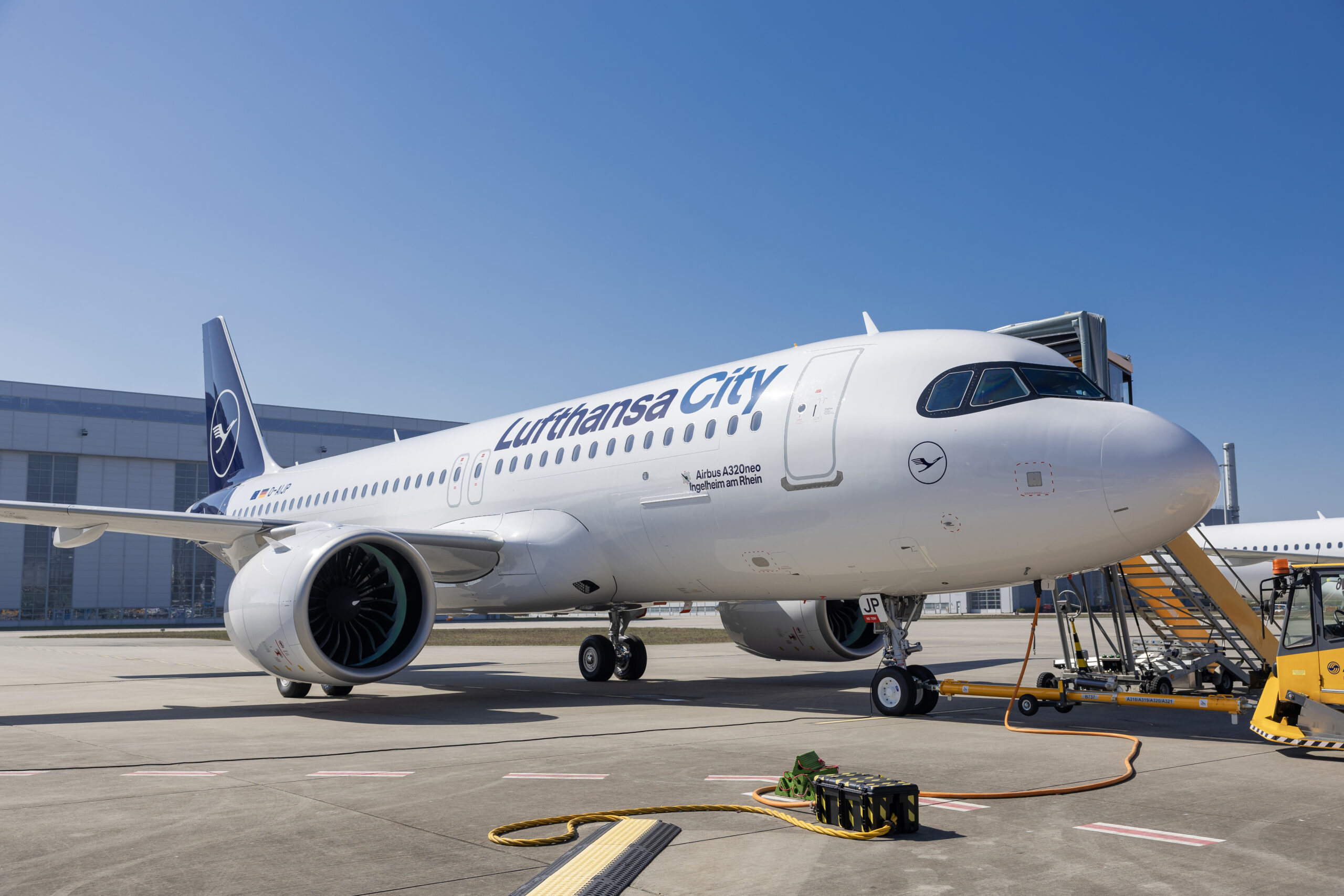 A Lufthansa City Airlines Airbus A320neo parked at an airport gate, connected to ground equipment under a clear sky.