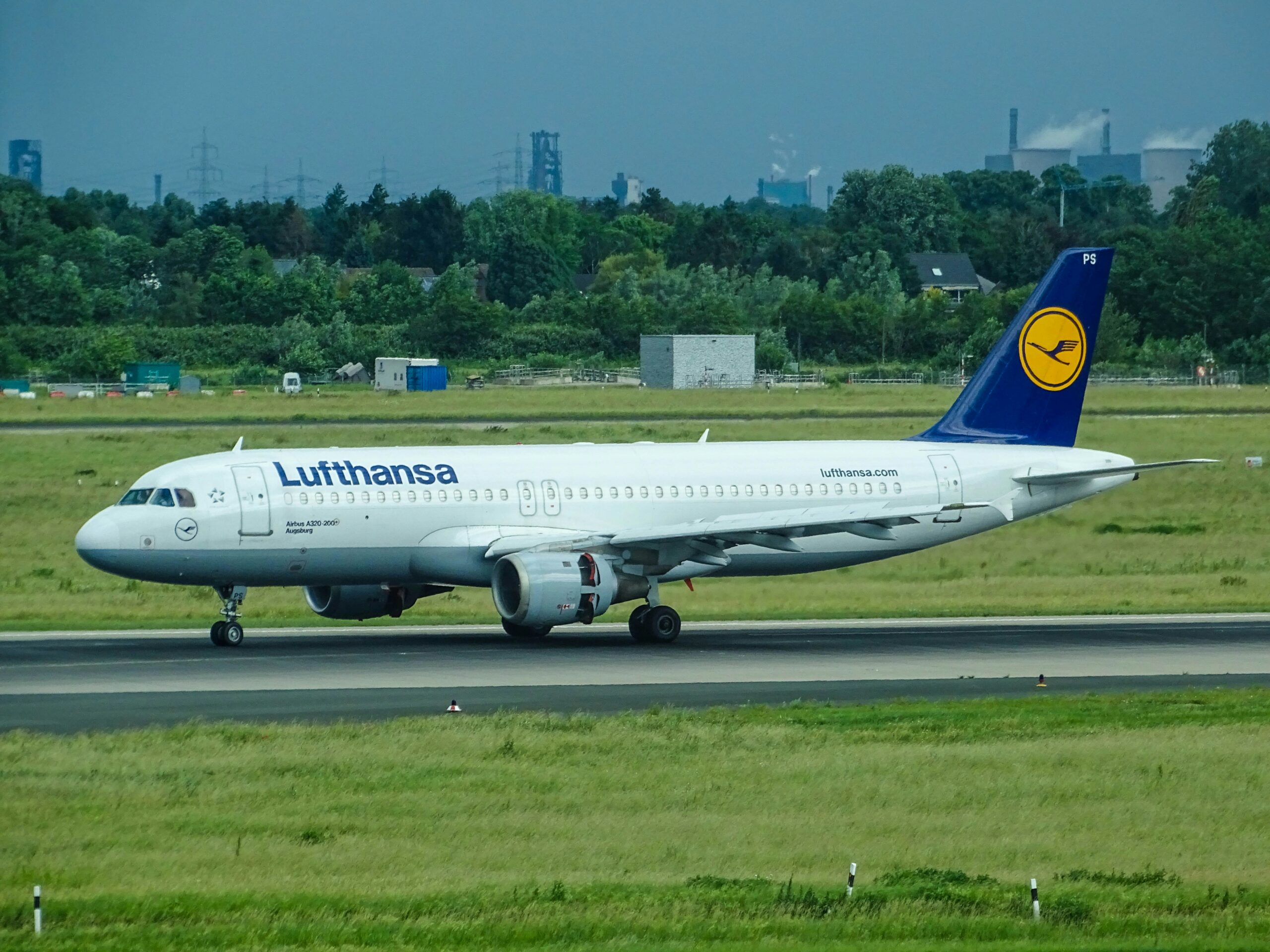 A Lufthansa aeroplane taxis on a runway, surrounded by lush greenery and industrial structures in the background.