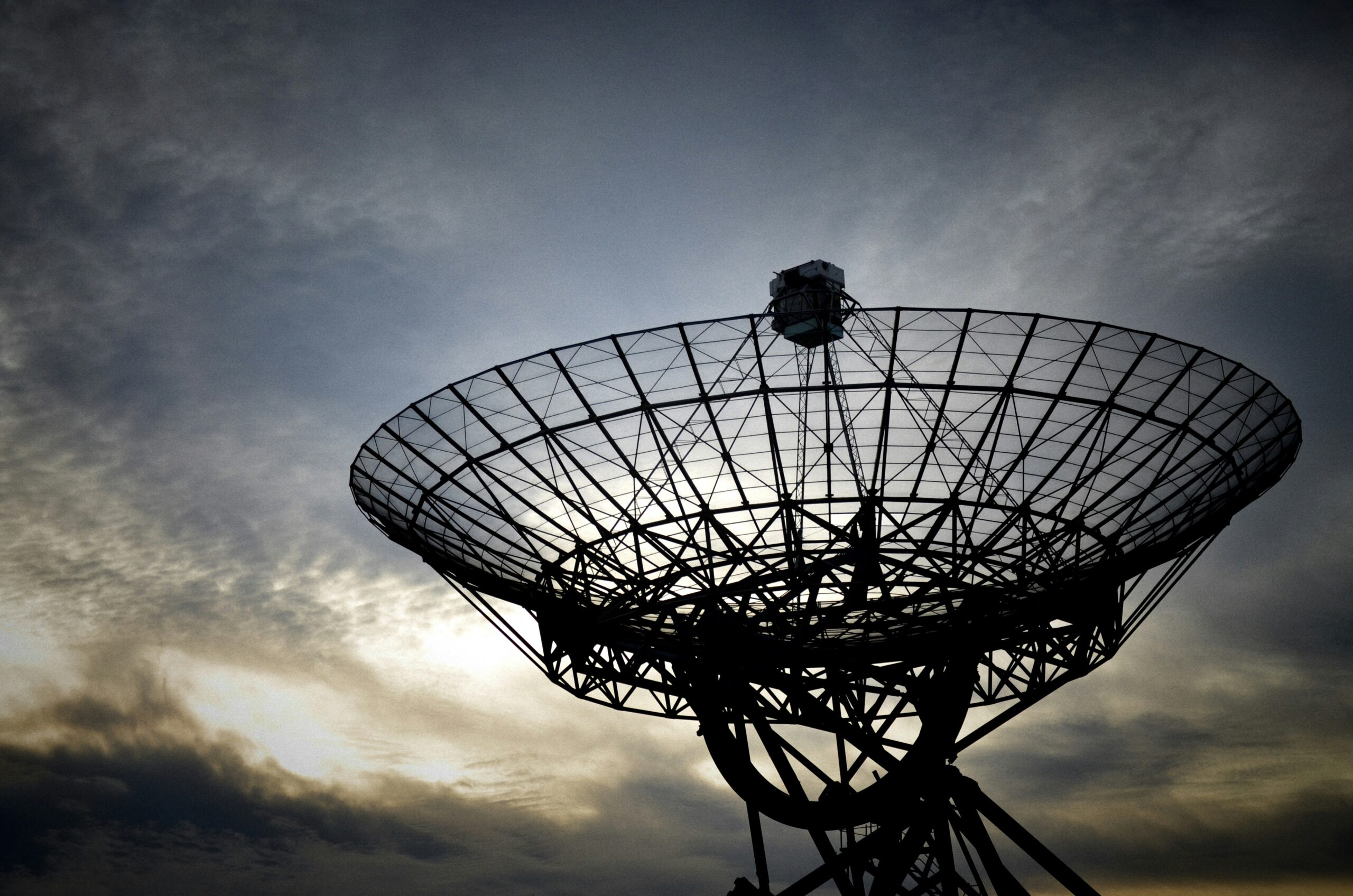 Silhouette of a satellite dish against a cloudy sky during sunset, representing space communication infrastructure.