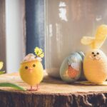 A charming Easter-themed setup featuring a small plush yellow chick wearing a hat adorned with flowers, a daffodil flower on the left, and a yellow yarn-wrapped bunny figure with floppy felt ears on the right. Behind them is a painted egg with a cute bunny illustration, all arranged on a rustic wooden surface.