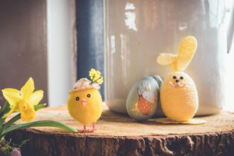 A charming Easter-themed setup featuring a small plush yellow chick wearing a hat adorned with flowers, a daffodil flower on the left, and a yellow yarn-wrapped bunny figure with floppy felt ears on the right. Behind them is a painted egg with a cute bunny illustration, all arranged on a rustic wooden surface.