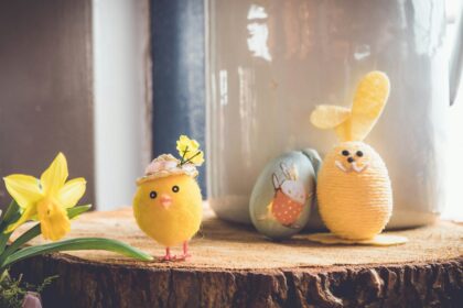 A charming Easter-themed setup featuring a small plush yellow chick wearing a hat adorned with flowers, a daffodil flower on the left, and a yellow yarn-wrapped bunny figure with floppy felt ears on the right. Behind them is a painted egg with a cute bunny illustration, all arranged on a rustic wooden surface.