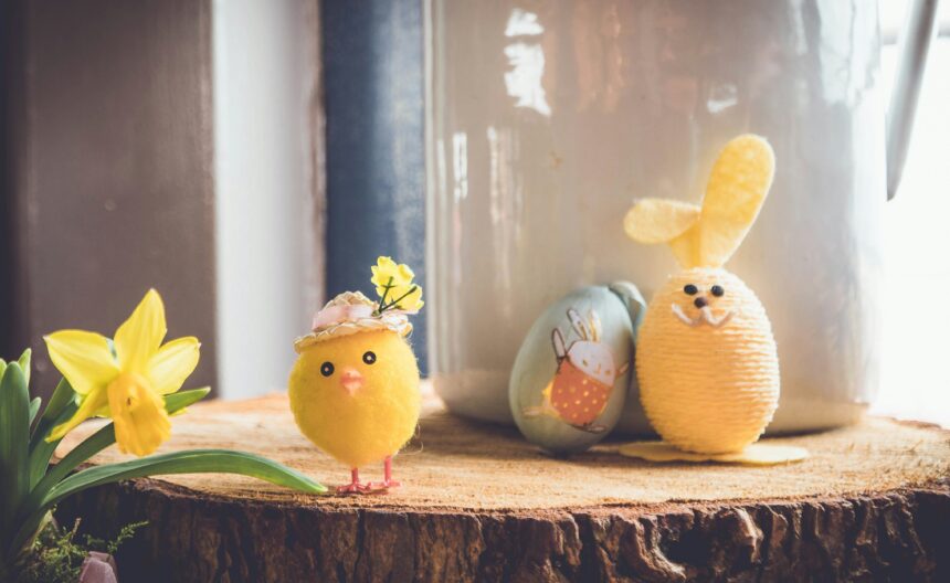 A charming Easter-themed setup featuring a small plush yellow chick wearing a hat adorned with flowers, a daffodil flower on the left, and a yellow yarn-wrapped bunny figure with floppy felt ears on the right. Behind them is a painted egg with a cute bunny illustration, all arranged on a rustic wooden surface.