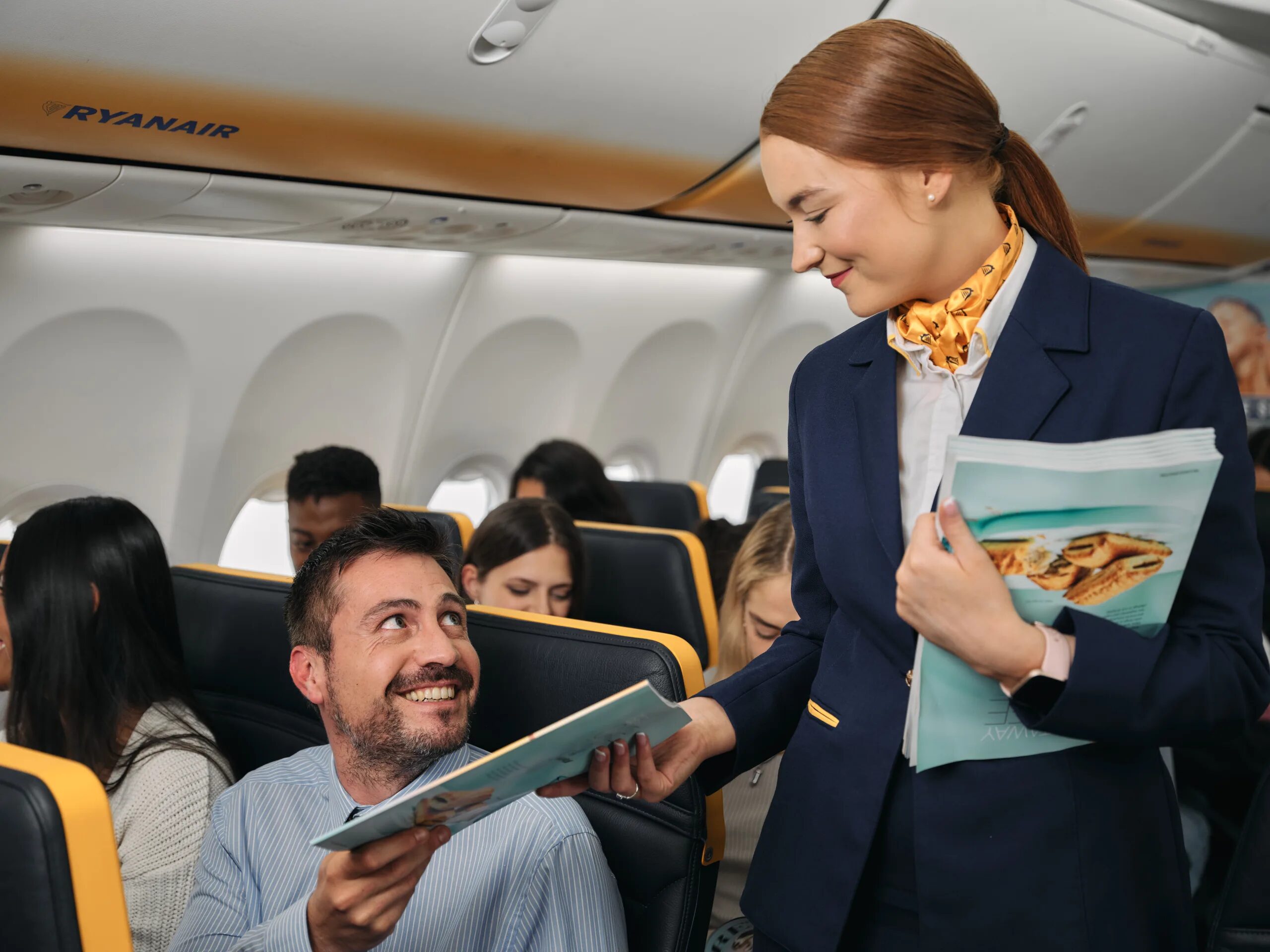 A happy flight attendant hands a passenger a magazine onboard an aircraft.