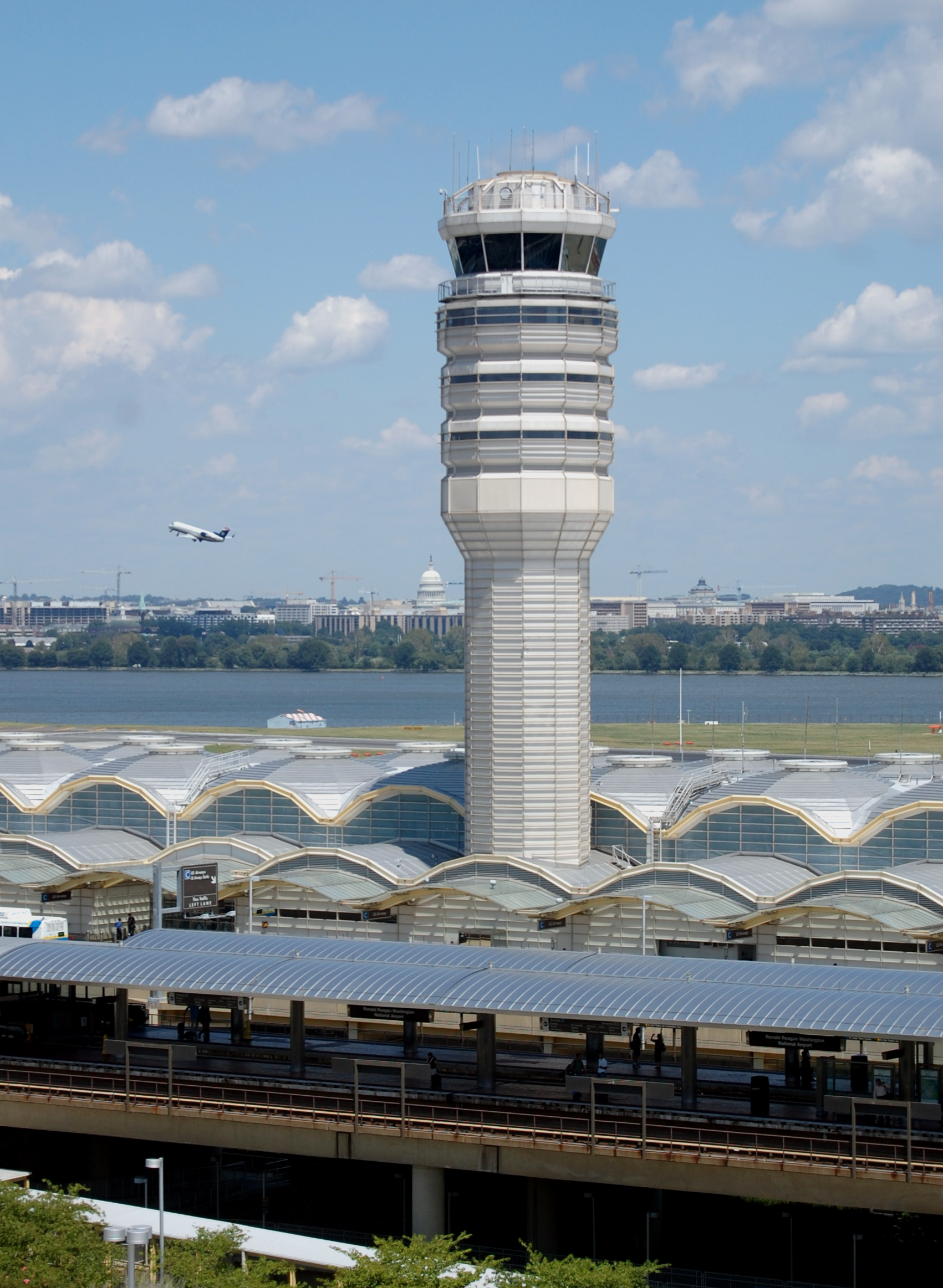 The tower and exterior of Ronald Reagan Washington National Airport. A plane is seen flying in the background.