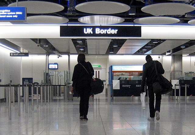 Two passengers walk towards a sign labelled 'UK Border' within an airport.