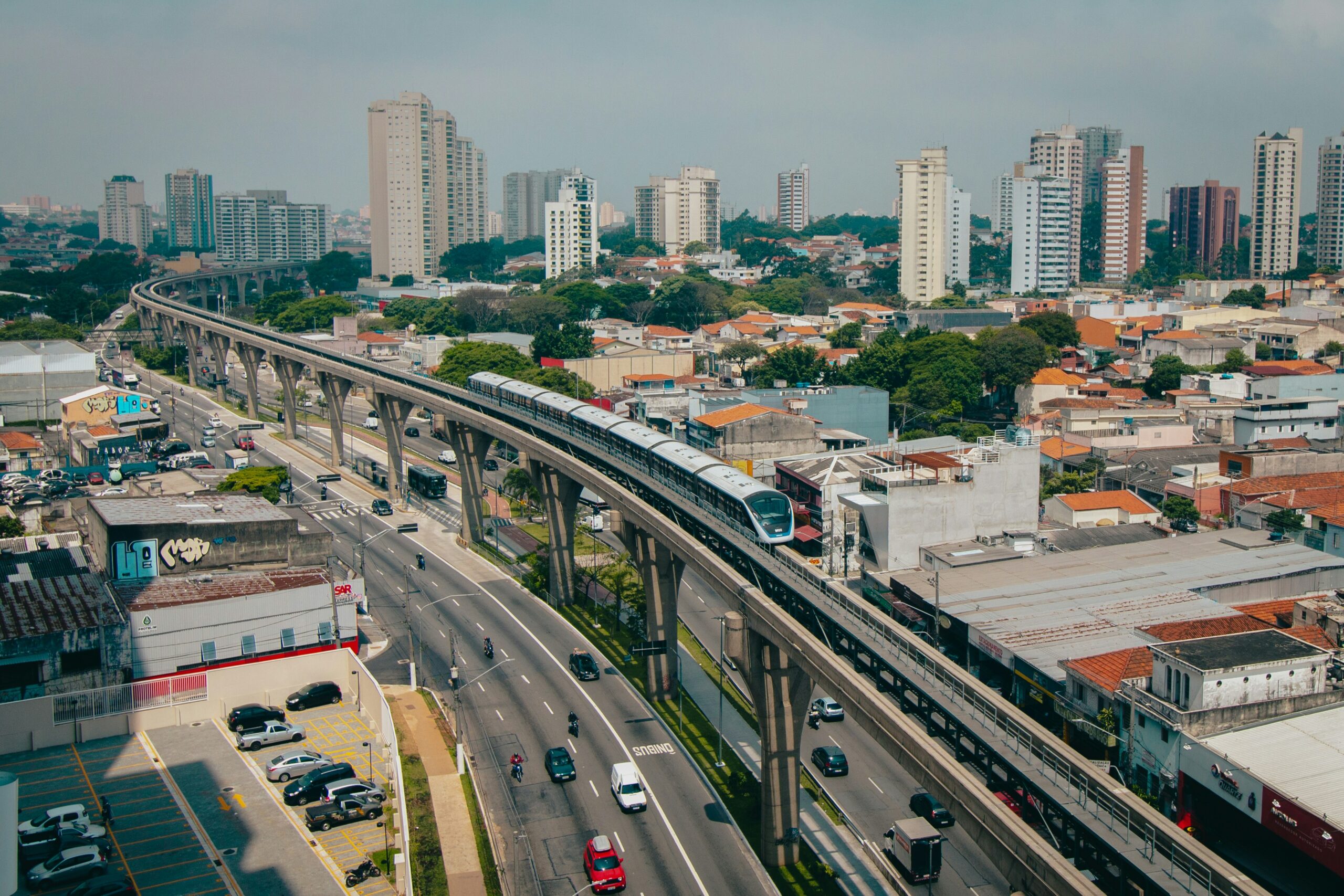 Aerial view of an urban area with a modern elevated train navigating through tall buildings. Busy streets with vehicles and greenery contrast against the cityscape.