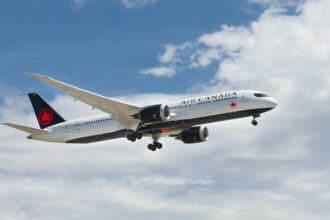 Air Canada airplane in mid-air with landing gear extended, flying through partly cloudy skies.