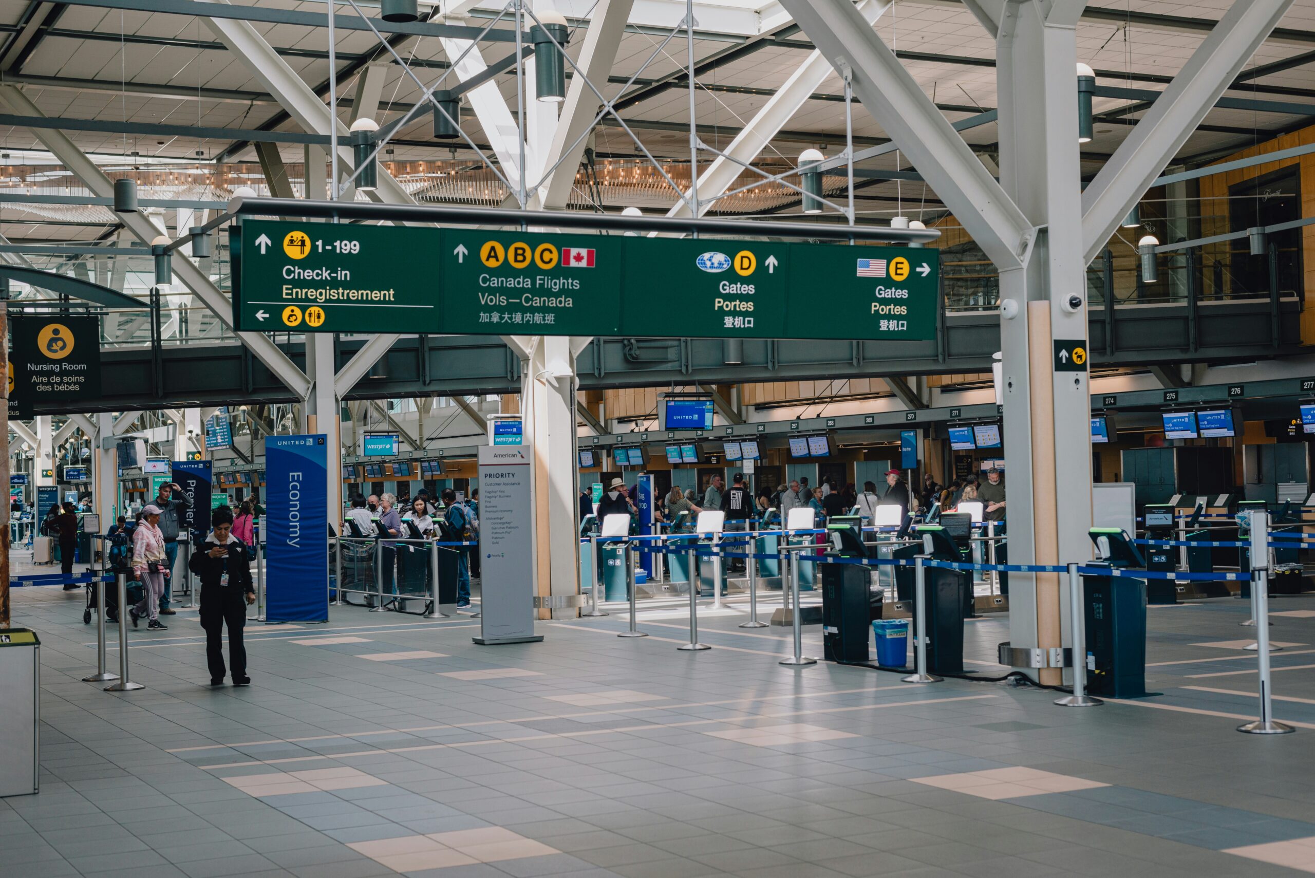 Interior view of an airport terminal with multiple check-in counters, digital screens, and passengers lining up; signage indicates directions for Canada flights and gates.