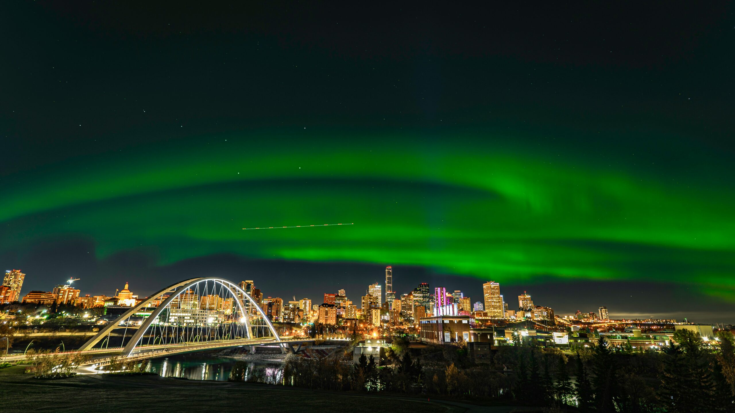 A vibrant green aurora illuminates the night sky over a city skyline, with a bridge and river in the foreground in Edmonton.