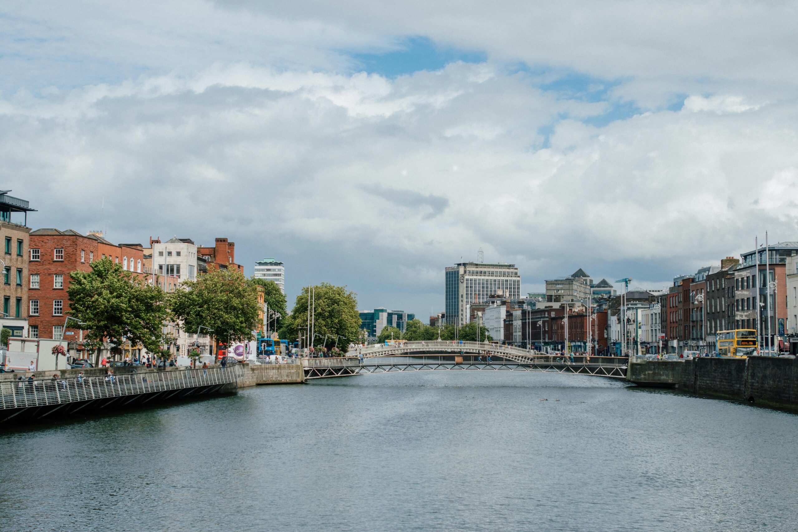 Urban riverside scene in Dublin, Ireland, featuring the River Liffey, Ha'penny Bridge, historic red-brick buildings, and people walking along the quay.