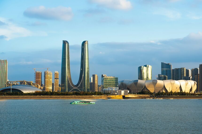 Waterfront view of Hangzhou skyline, showcasing modern architecture with futuristic skyscrapers and the distinctive Lotus-Flower-Shaped building under a clear blue sky, reflecting in the river.